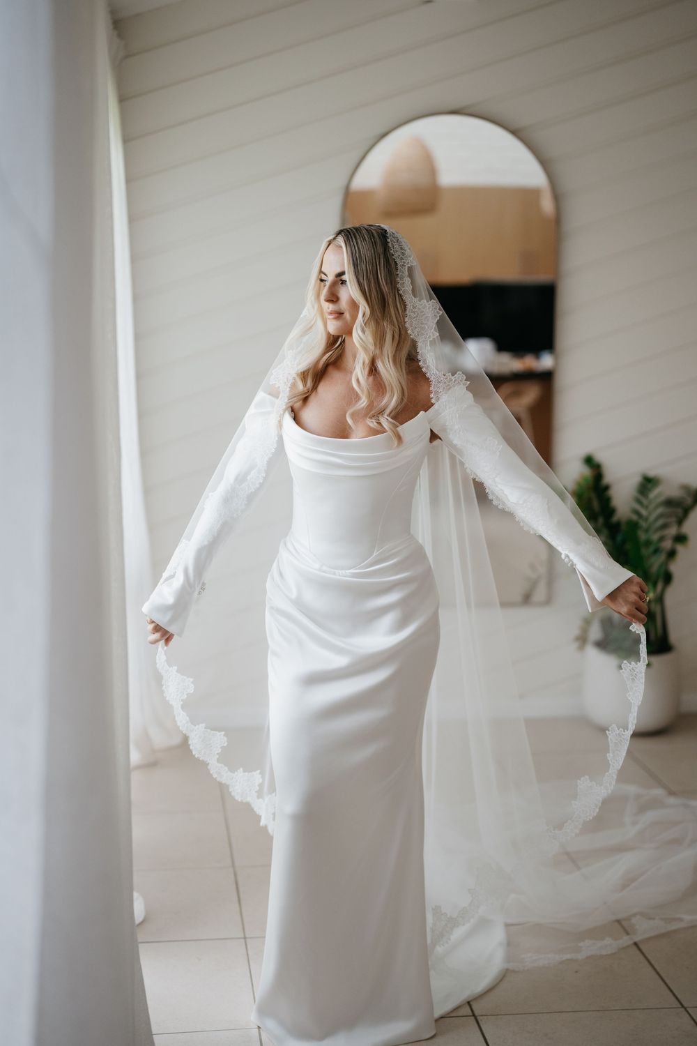 Elegant bride in form-fitting white wedding dress with flowing veil poses gracefully in arched doorway.