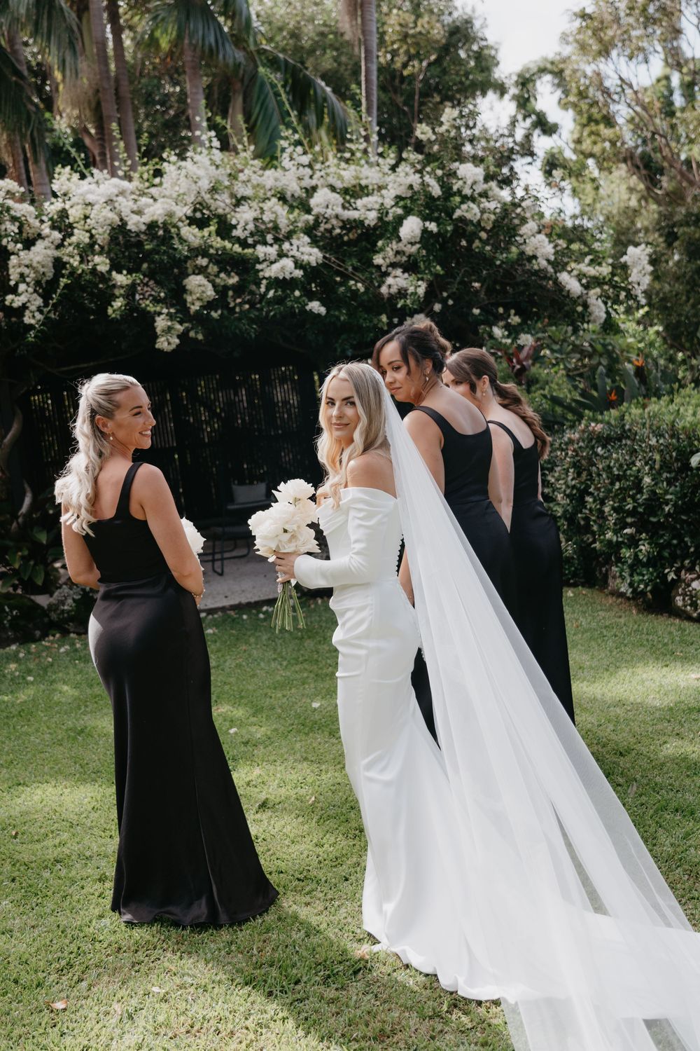 Wedding party stands together on a sunny lawn while bridesmaids in black dresses arrange the bride's flowing veil.