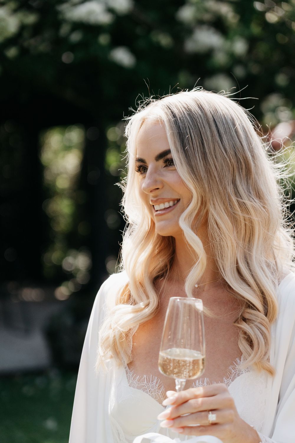 Blonde bride in wedding dress smiles while holding champagne glass outdoors in natural sunlight.