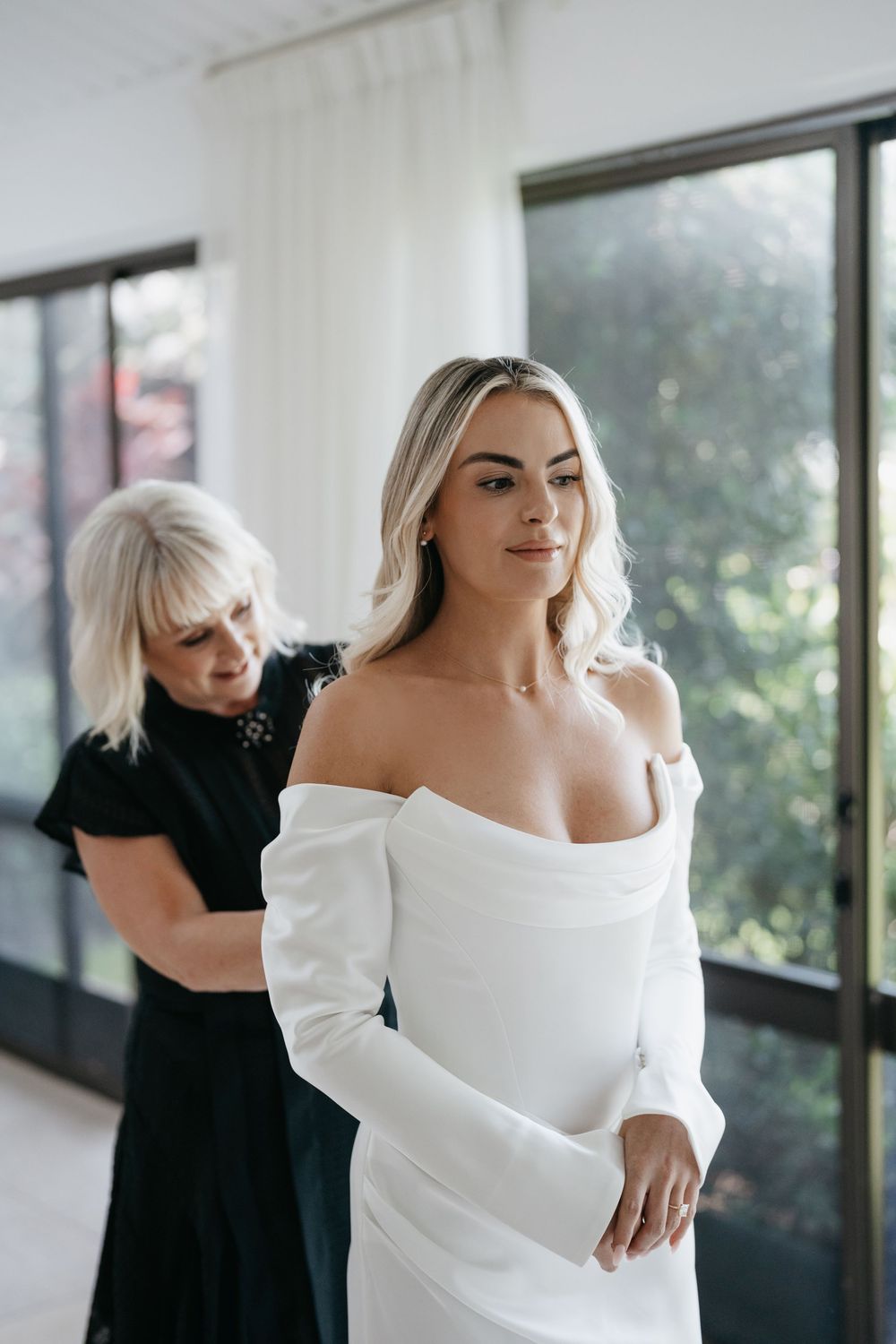 Elegant white off-shoulder wedding dress being styled in front of bright windows.