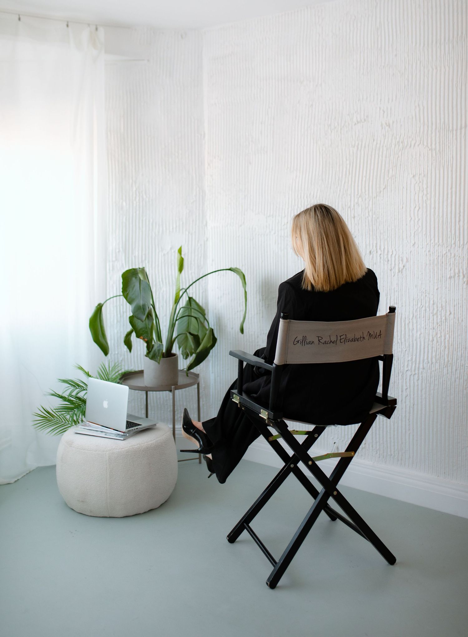 Black director's chair positioned against white textured wall with potted plants creating minimalist indoor scene.
