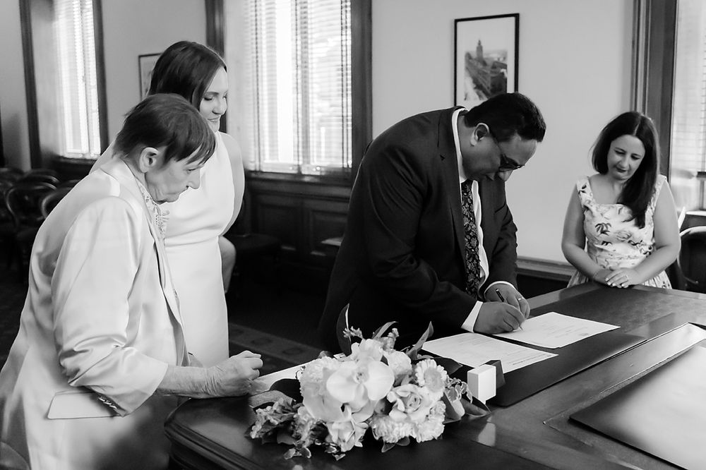 Black and white image of an intimate courthouse ceremony with people gathered around paperwork and flowers on the desk.