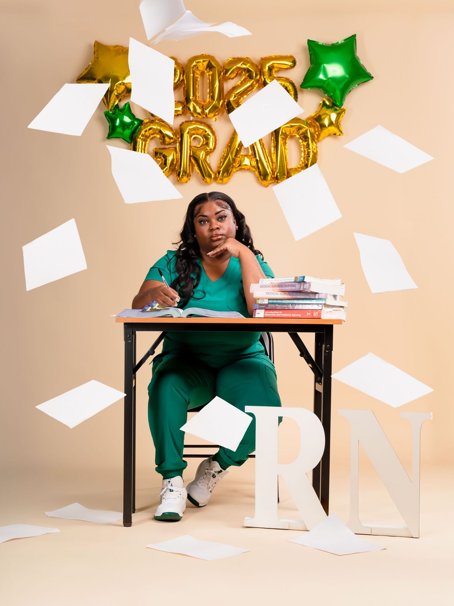 Creative graduation photo shoot with flying papers and gold letter balloons against a beige backdrop.