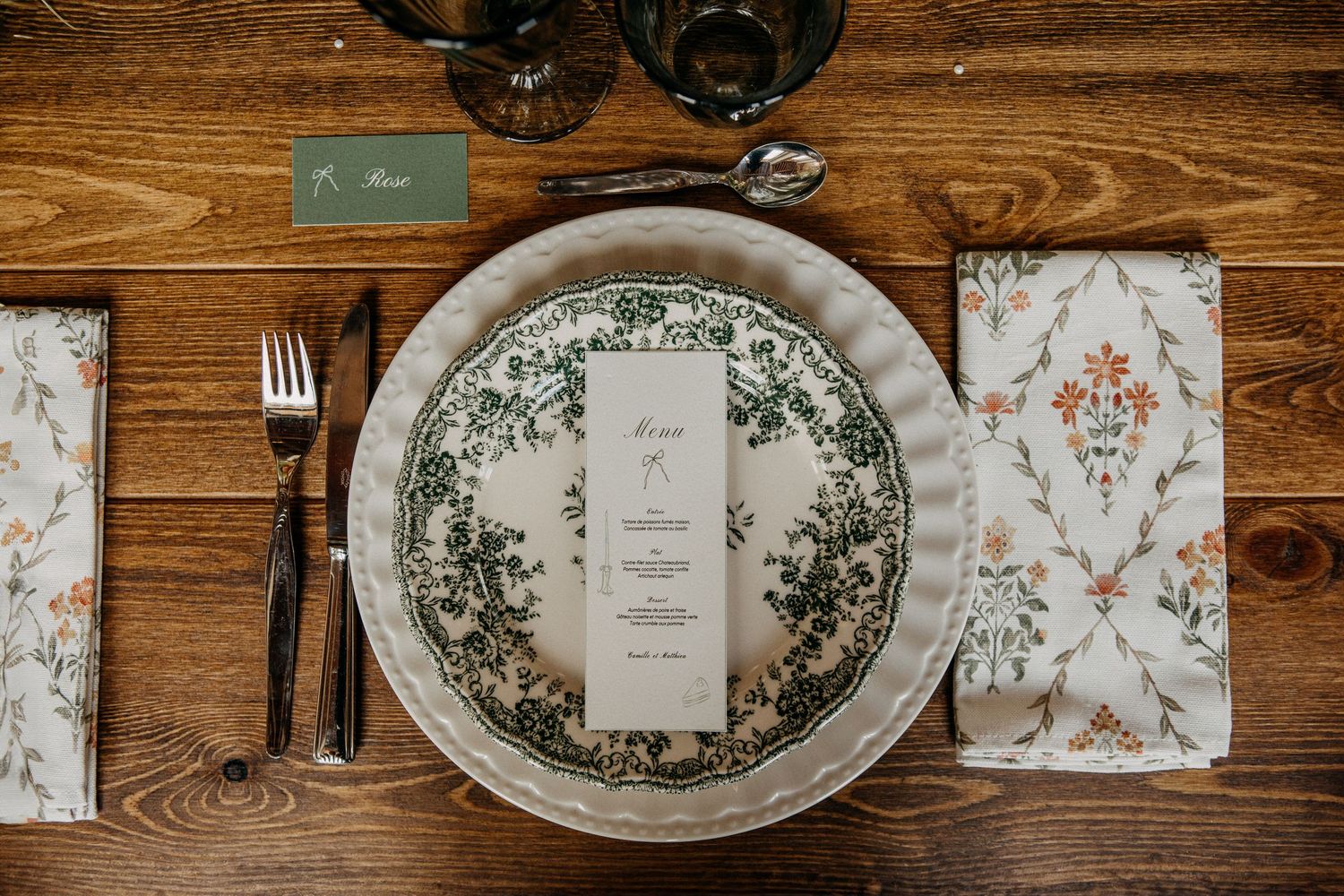 Elegant place setting with green floral china plate, menu card, patterned napkin, and black cutlery on wood.