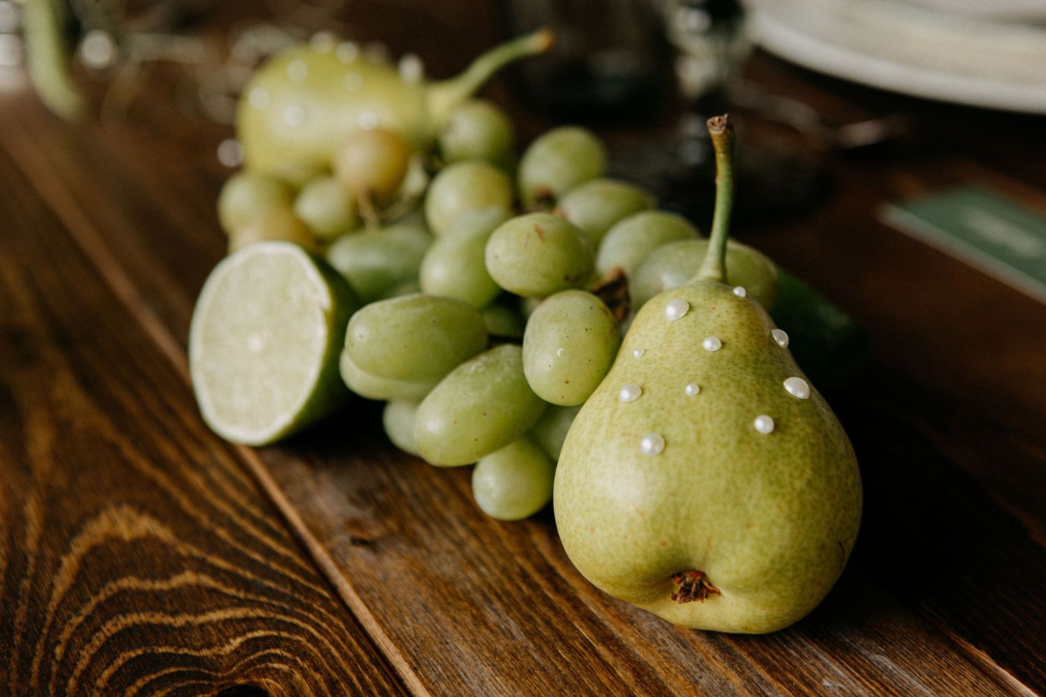 Fresh green grapes, pear with pearl accents, and halved lime arranged on a dark wooden table.
