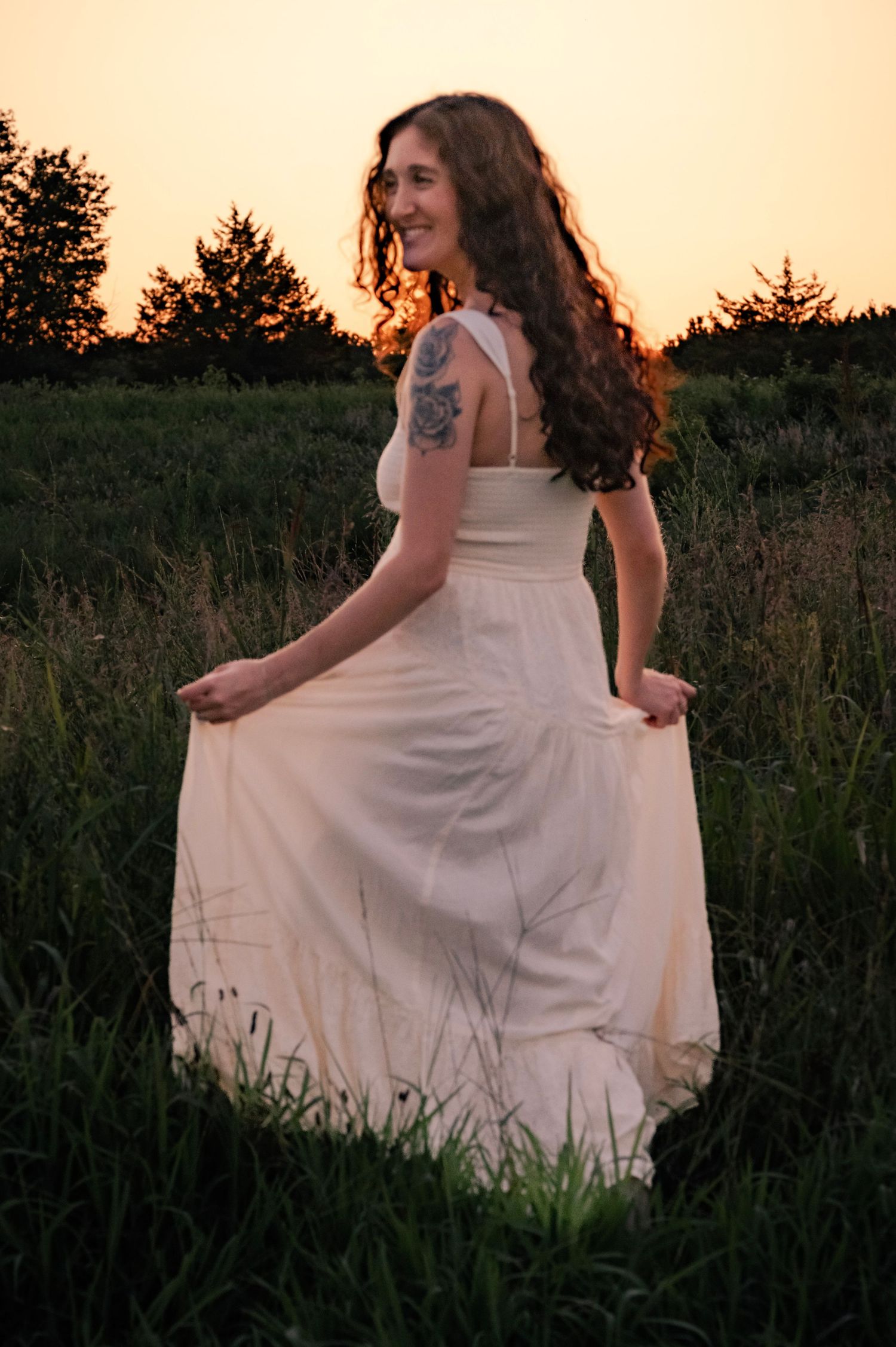 Someone wearing a white flowy dress twirls gently in a meadow at golden hour sunset, surrounded by tall grass.