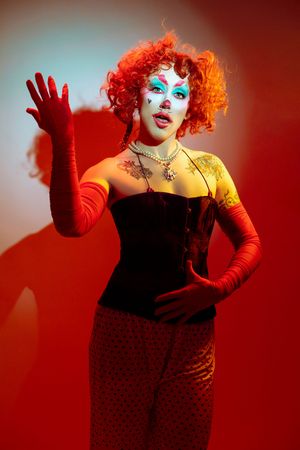 Drag performer in black corset and red gloves poses dramatically with bright orange curly hair against red backdrop.
