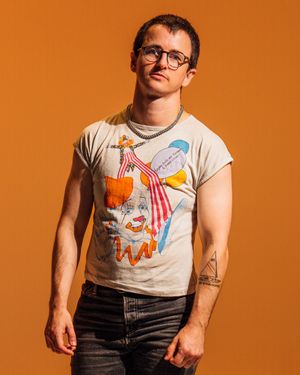 Person in glasses and graphic t-shirt with colorful design poses against orange backdrop in studio setting.