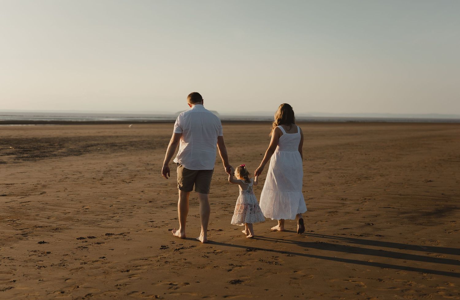 A couple and young child walk along a beach at sunset holding hands, casting long shadows on the sand.
