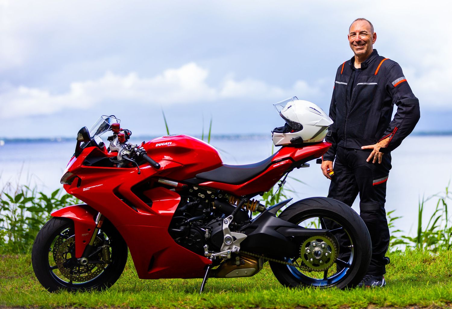 A bright red sportbike motorcycle parked on green grass near the water with someone standing beside it.