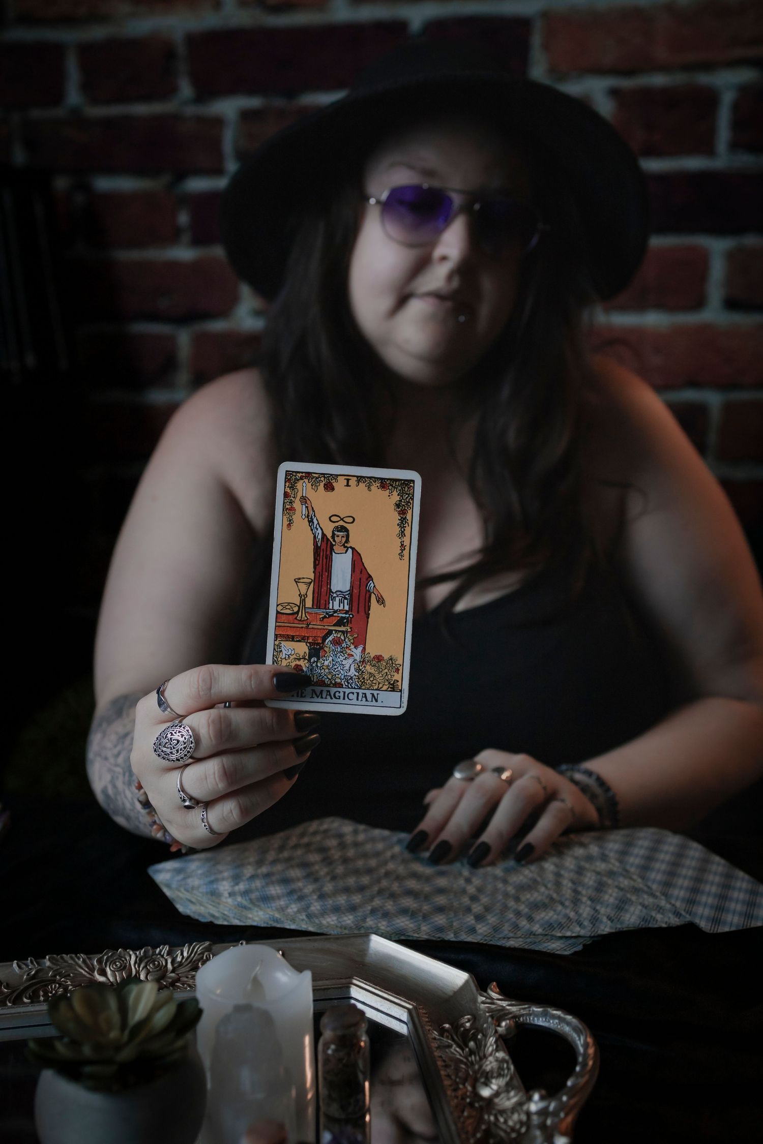 A fortune teller holds up the King of Wands tarot card while wearing a black hat and purple glasses at a brick wall setting.