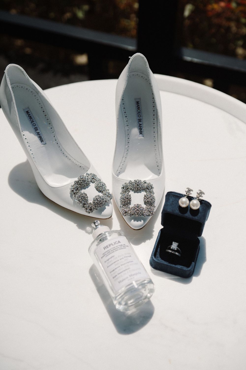 Elegant white bridal shoes with crystal embellishments displayed next to jewelry box and water bottle in sunlight.