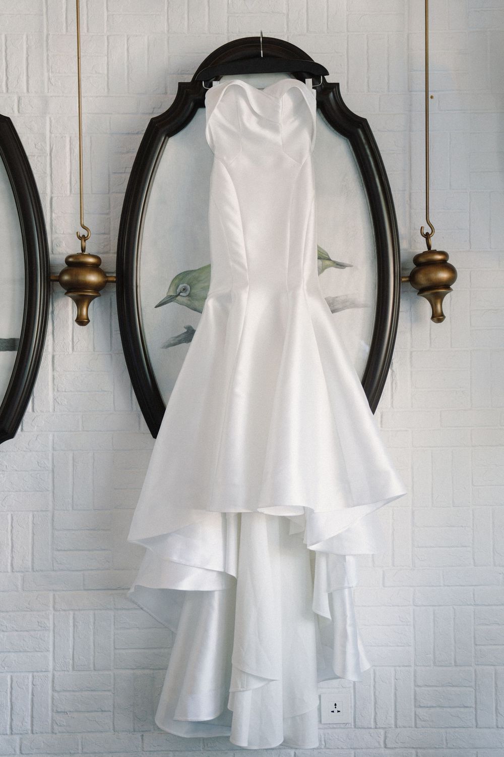 White wedding dress hangs elegantly on a black oval frame against a textured white wall with antique brass sconces.