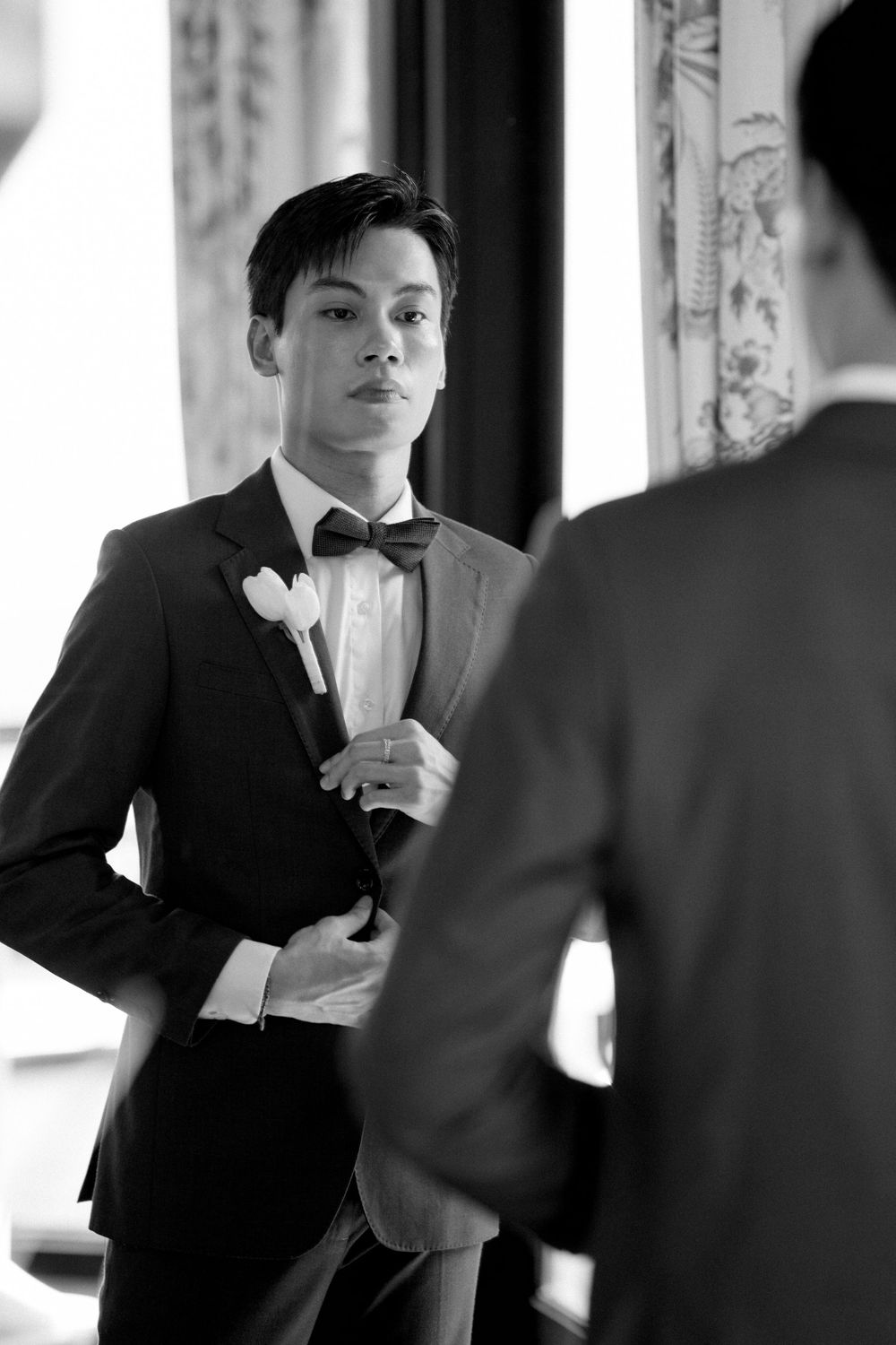 Black and white photo of someone in a formal tuxedo adjusting their bow tie while looking in a mirror.