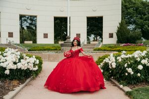 A stunning red quinceañera dress photographed against a white stone building with blooming flowers.