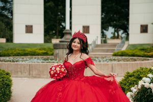 Elegant red ball gown with off-shoulder design photographed in an outdoor garden setting.