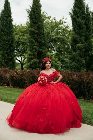 A stunning red quinceañera ball gown with floral details photographed in an outdoor garden setting.
