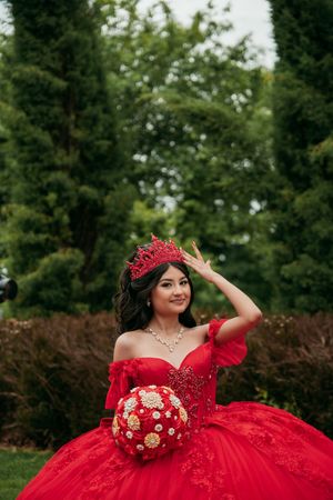 An elegant red formal dress with a crown accessory and floral bouquet in a garden setting.