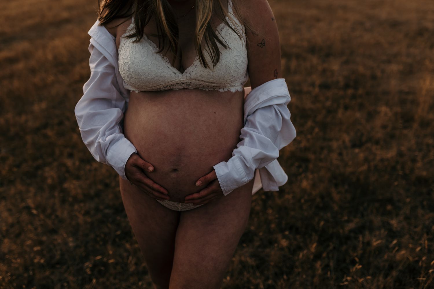 Pregnant woman in white top and tan underwear standing in field at sunset, touching belly in intimate maternity photo.