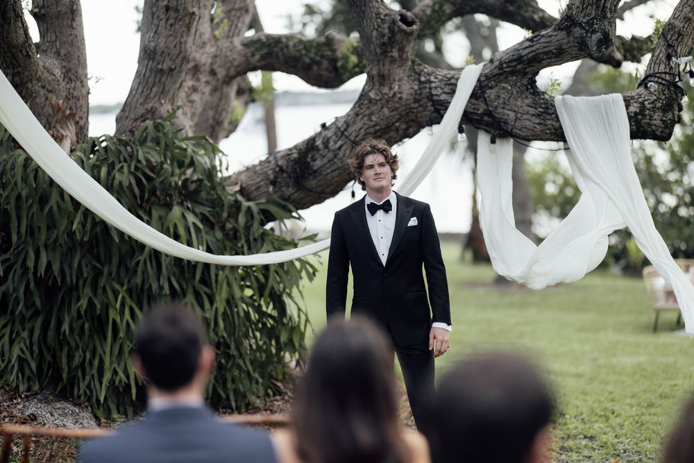 A groom stands under a large tree decorated with flowing white fabric during an outdoor wedding ceremony.