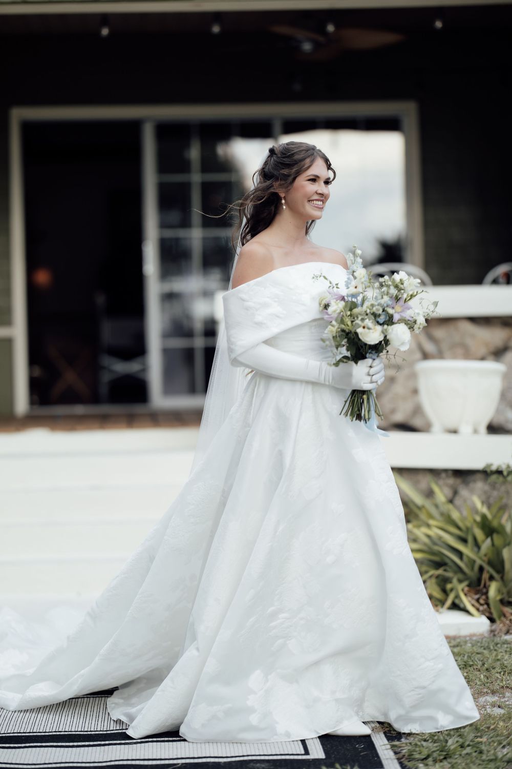 A flowing white wedding dress billows in the breeze as someone sits on outdoor steps with a floral bouquet.