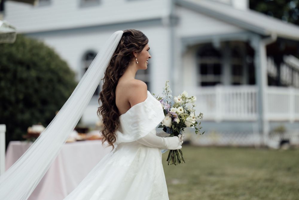A bride in a flowing white wedding gown holding a bouquet poses outdoors against a nature backdrop.