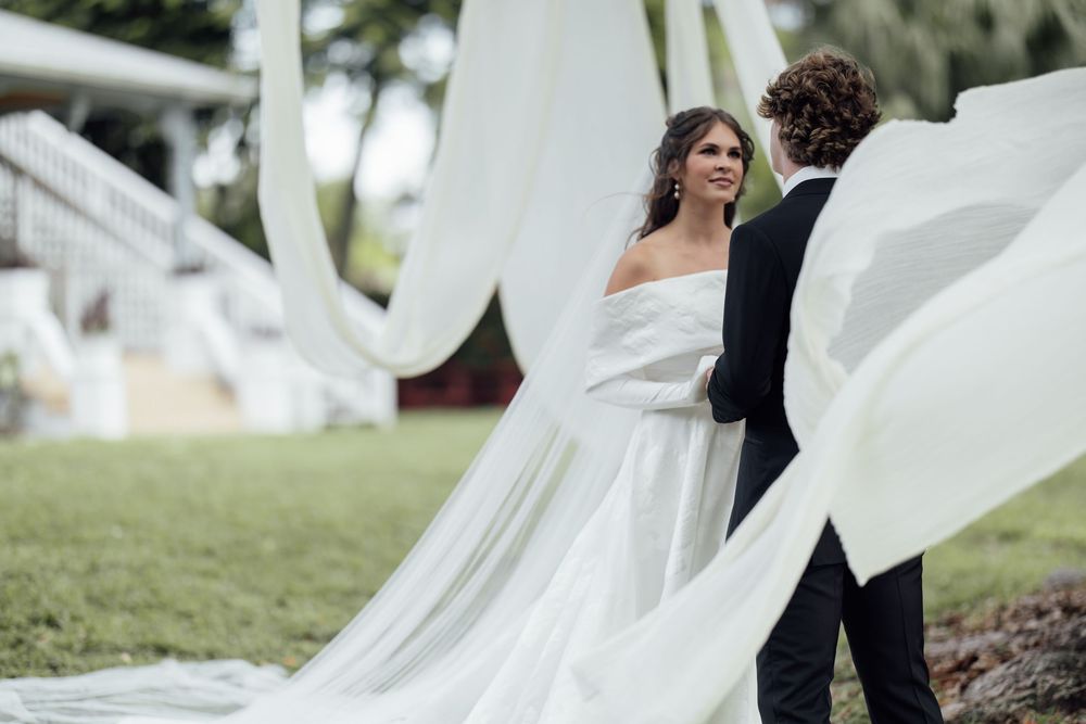 A flowing white wedding dress veil catches the wind during an outdoor photoshoot in a grassy setting.