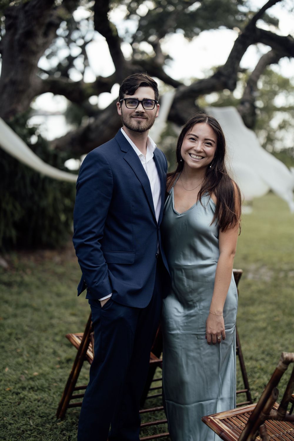 Stylish couple poses outdoors with vintage wooden chairs in a romantic garden photoshoot.