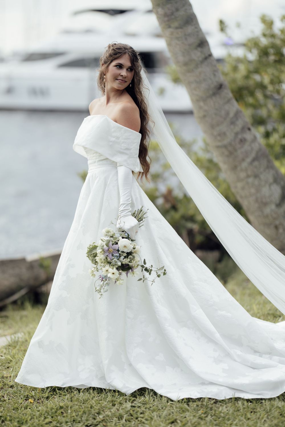A bride in an elegant off-shoulder wedding gown holds a delicate bouquet against a waterfront backdrop.