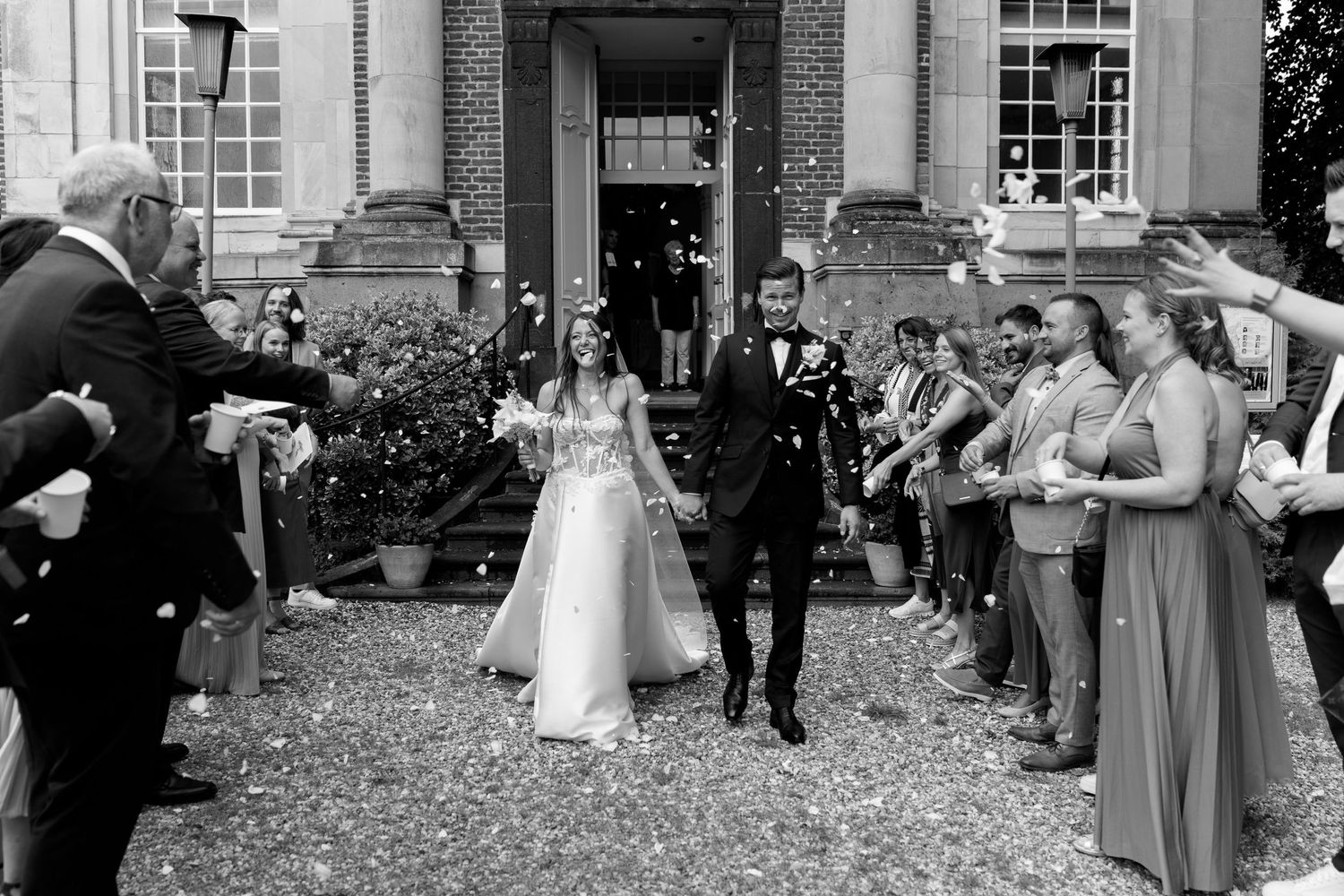 Wedding party exits venue as guests shower them with confetti in black and white.