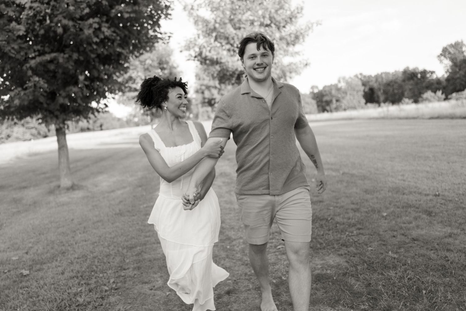 A joyful couple runs playfully through a grassy field on a sunny day during their photo shoot.