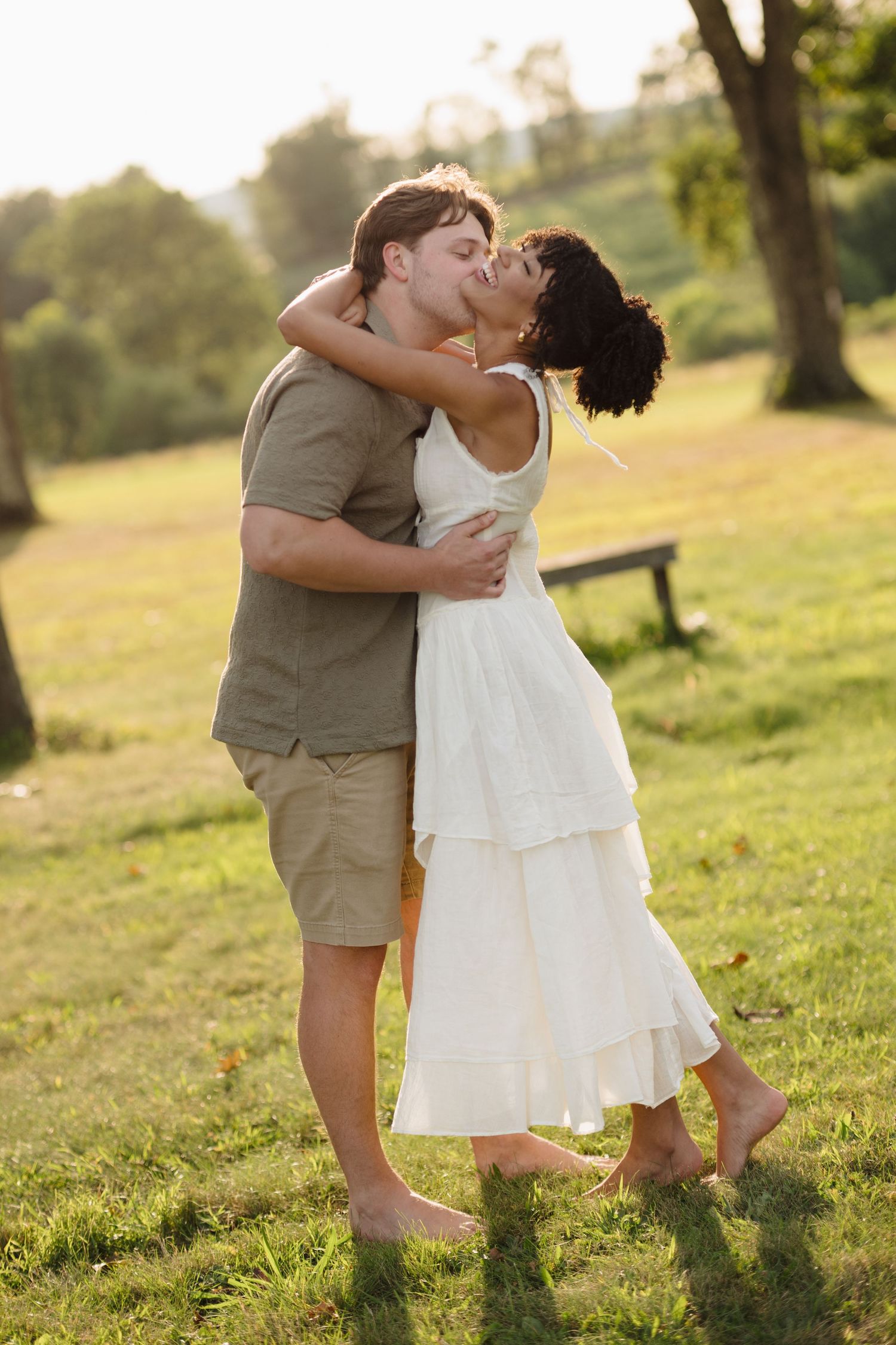 A romantic couple embraces and kisses in a sunlit park with green grass and trees in the background.