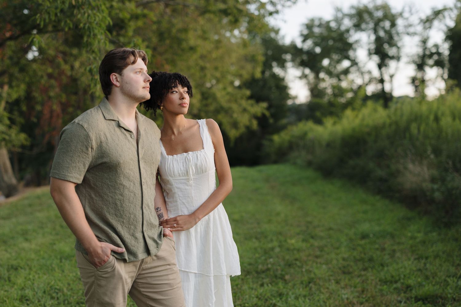 A couple stands together in casual summer attire against a lush green background.