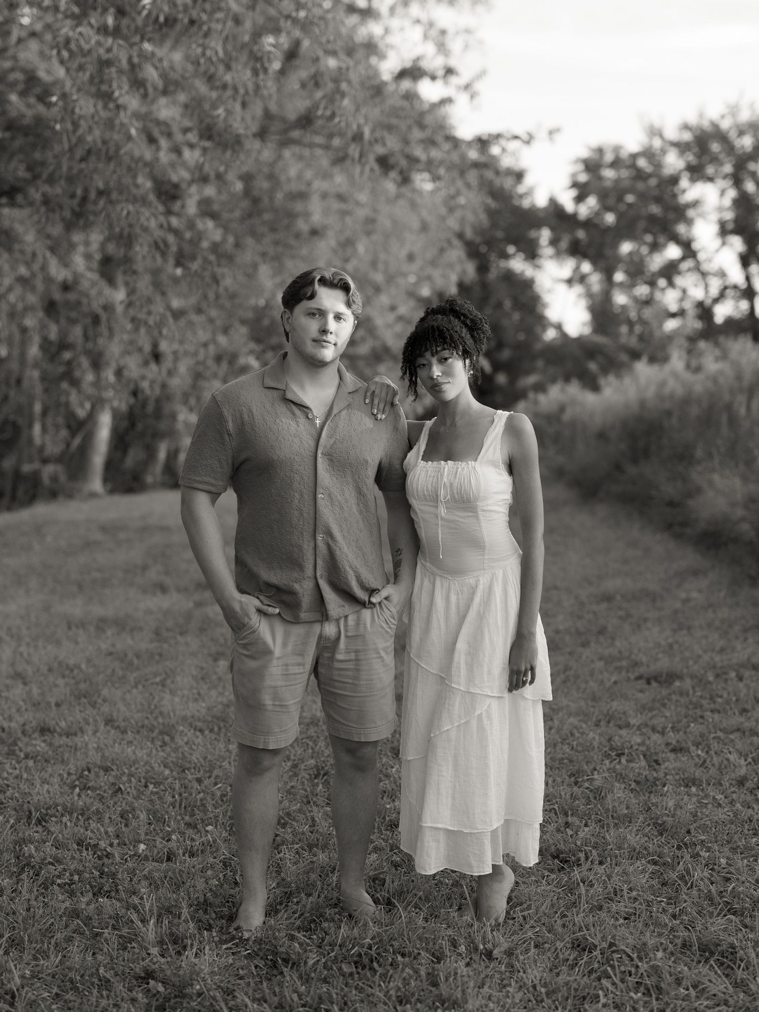 A black and white portrait of a couple standing together on a tree-lined path.