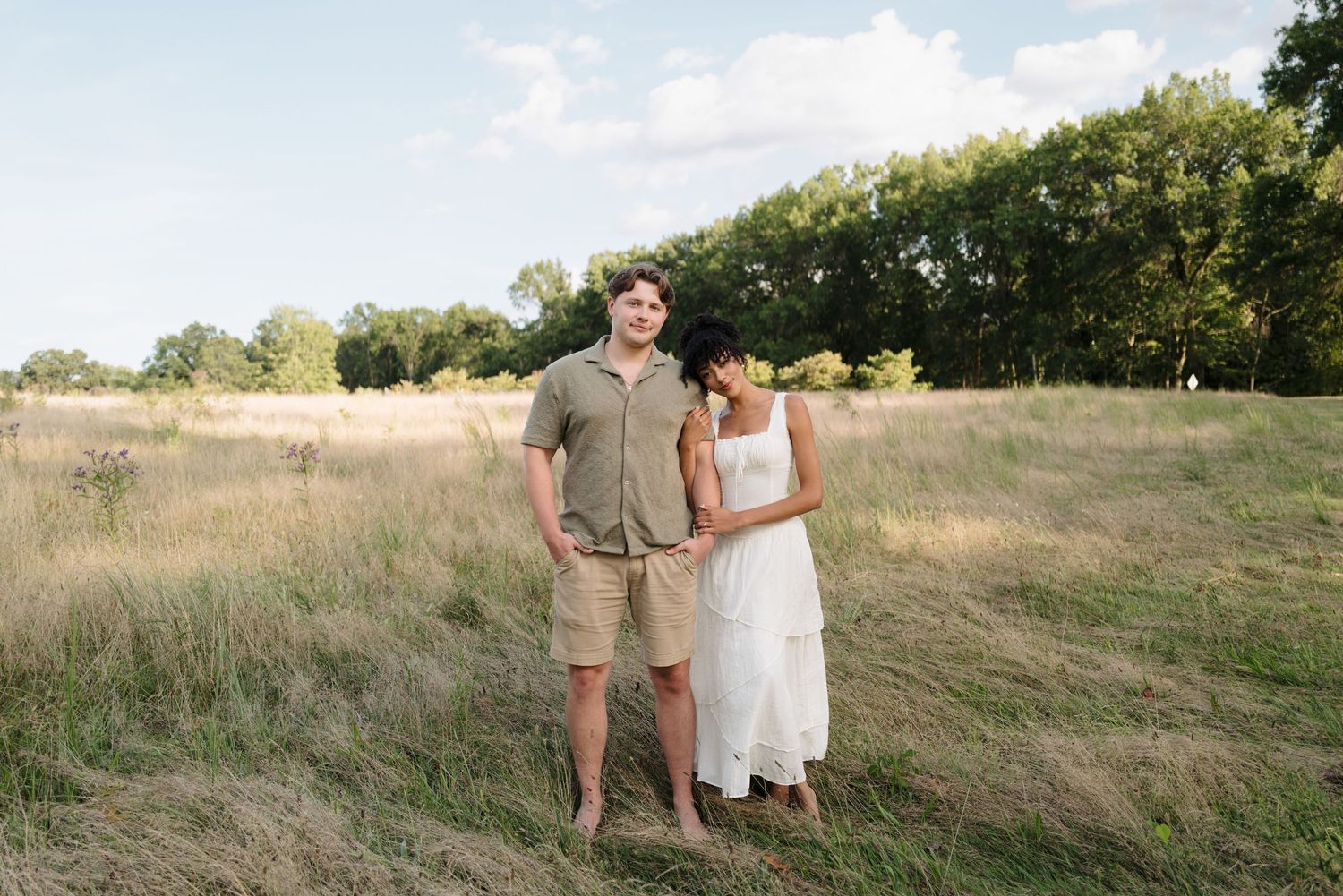 A couple poses together in a scenic field with rolling hills and blue sky in the background.
