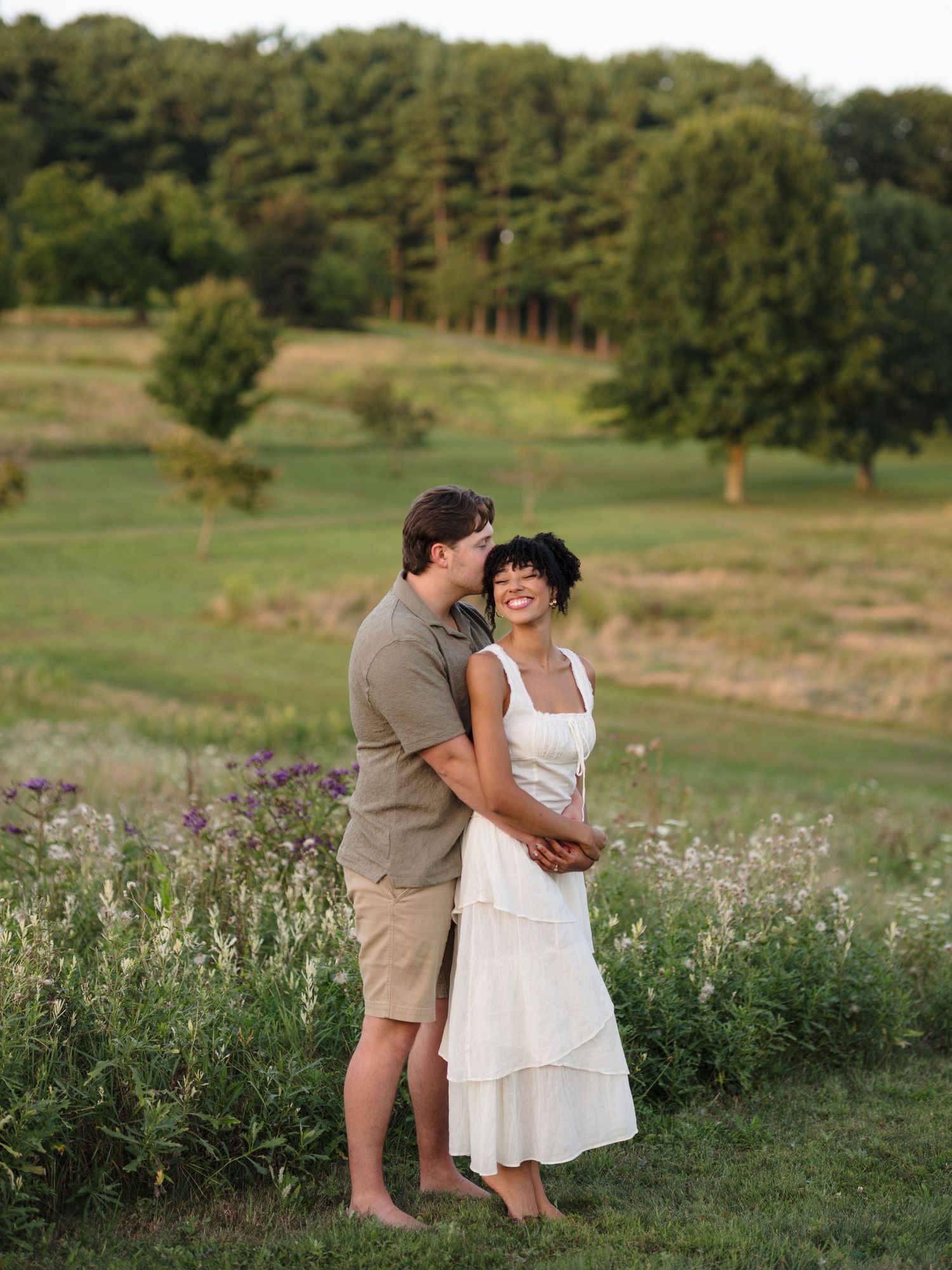 Two people share playful moments in a sunny field with wildflowers and trees in the background.