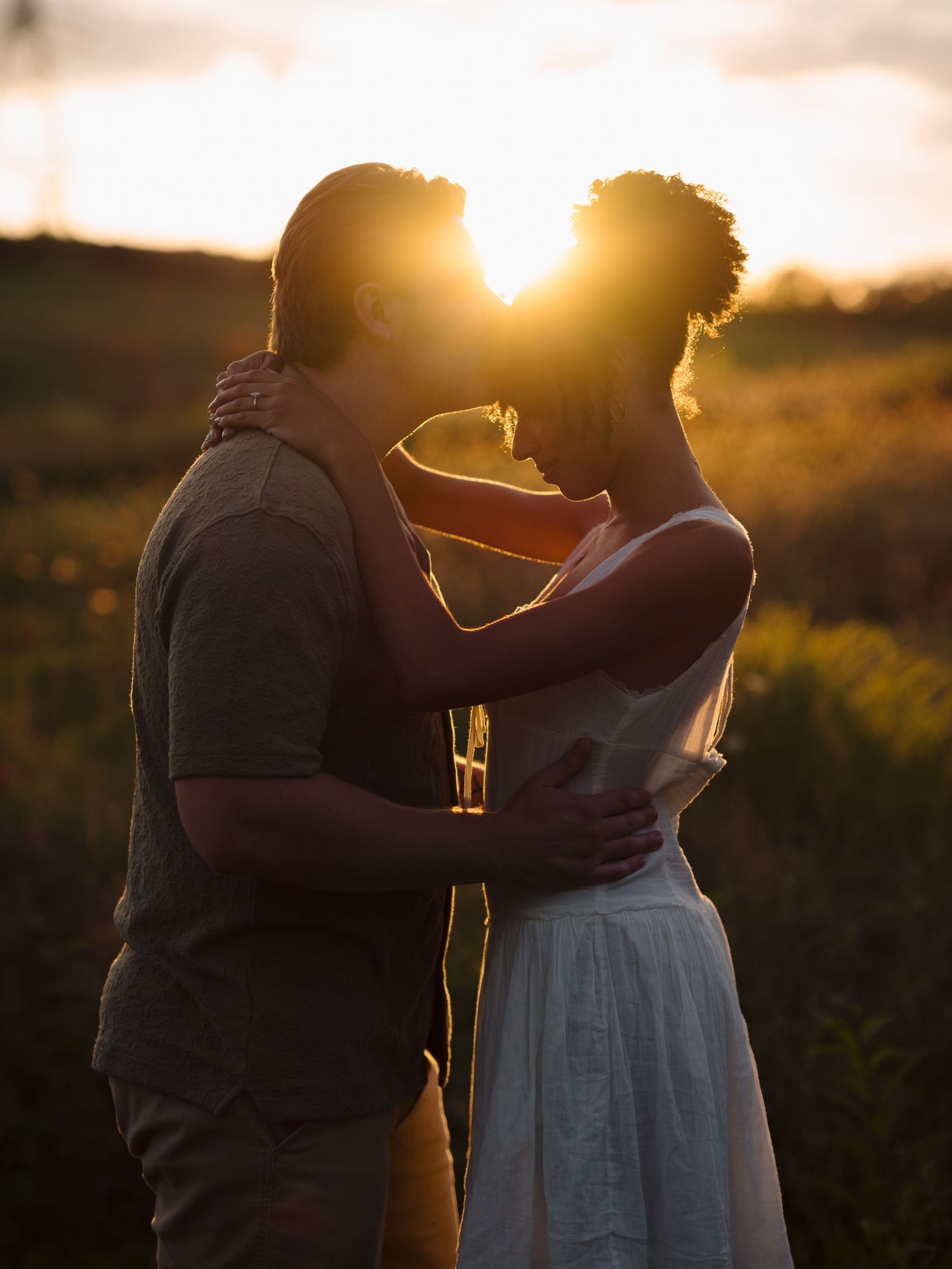 A couple embraces in silhouette against a glowing sunset backdrop in a natural outdoor setting.