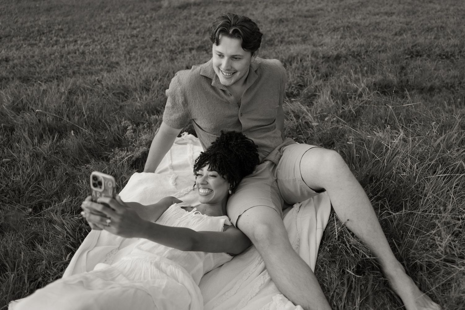 Black and white photo of a couple laying in grass with one person holding a phone while relaxing together.