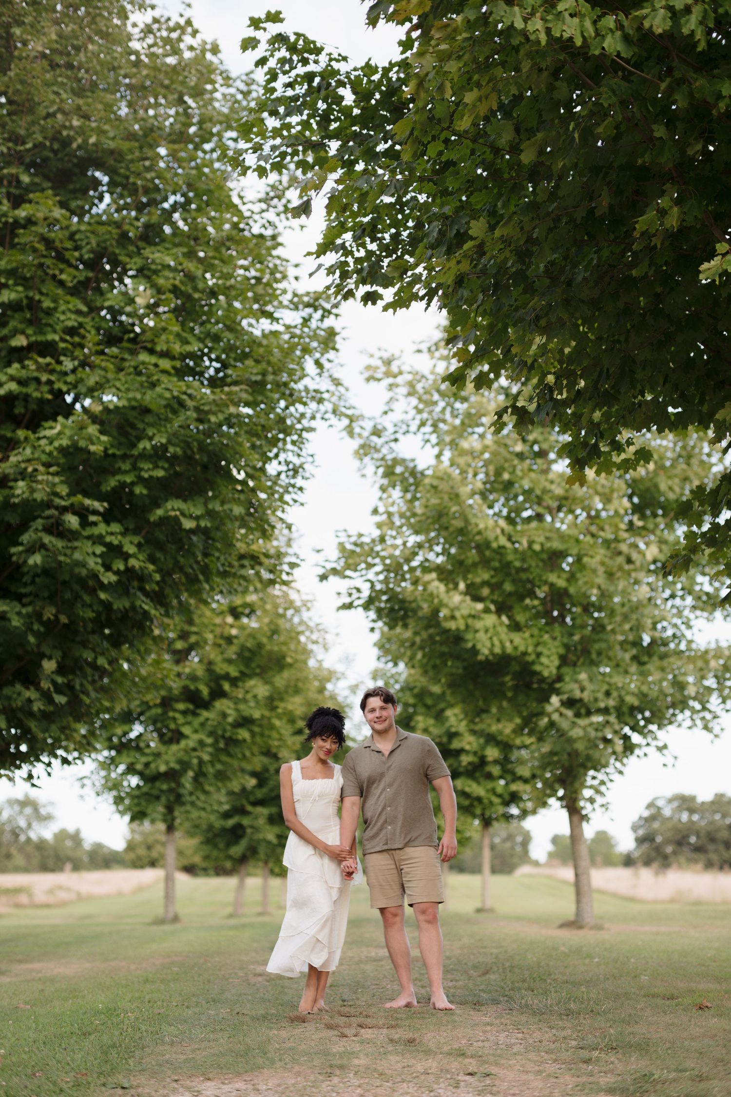A couple walks hand in hand beneath towering green trees in a peaceful park setting.