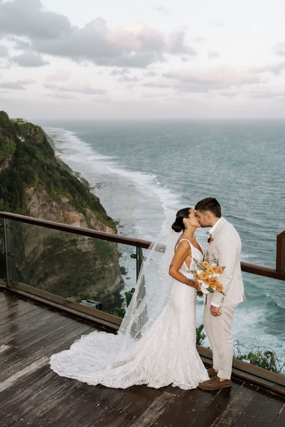 A romantic coastal wedding moment captured on a dramatic clifftop deck overlooking the ocean in misty conditions.