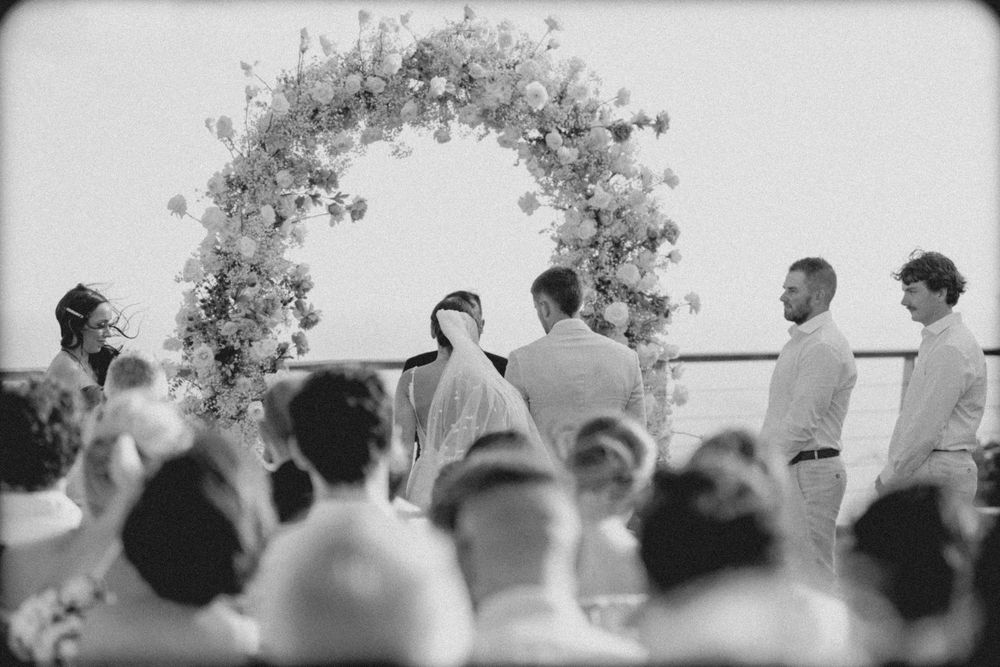 A classic black and white photograph shows a wedding ceremony taking place under a floral archway with guests seated.