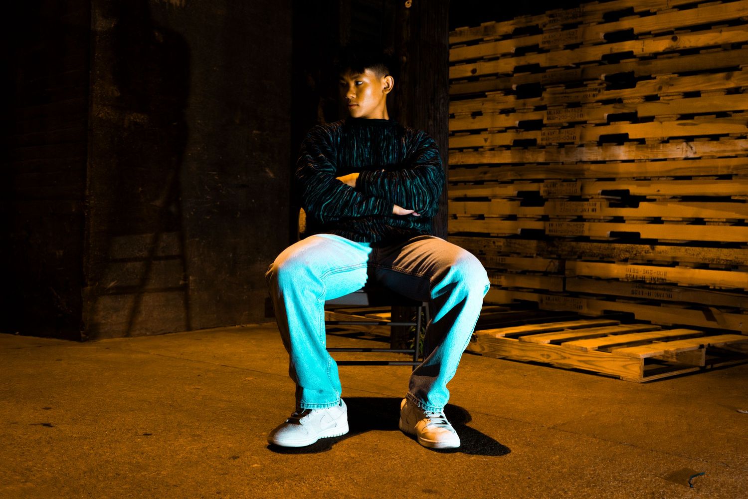 A person in a dark jacket and light blue pants sits on a wooden pallet in a dimly lit warehouse setting.
