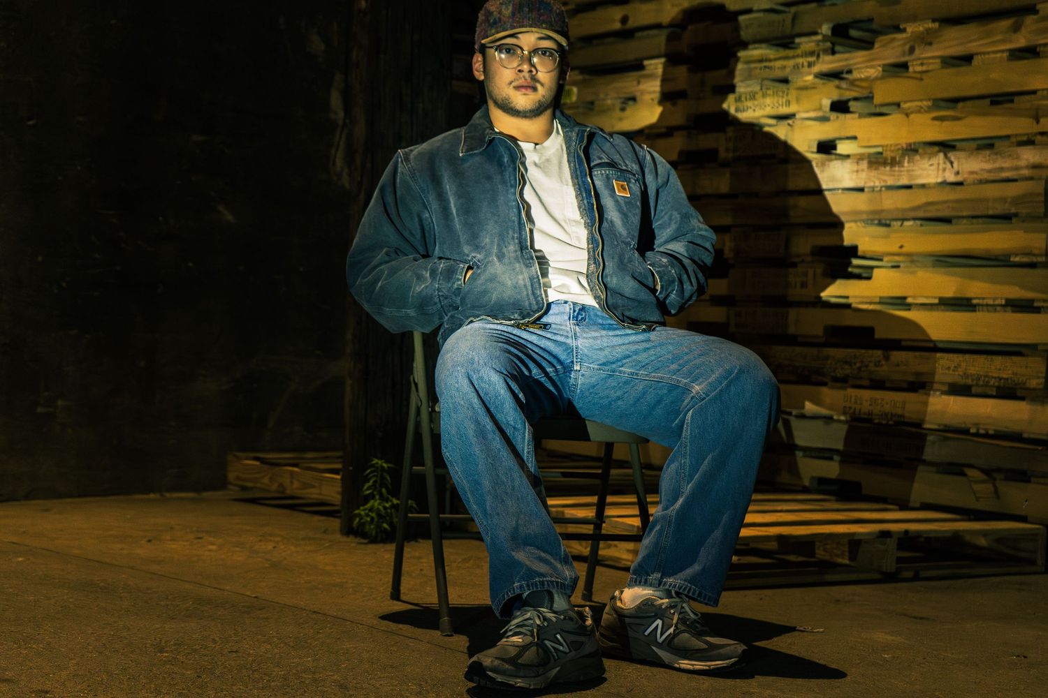 Person in denim jacket and jeans seated on chair in dramatic lighting.