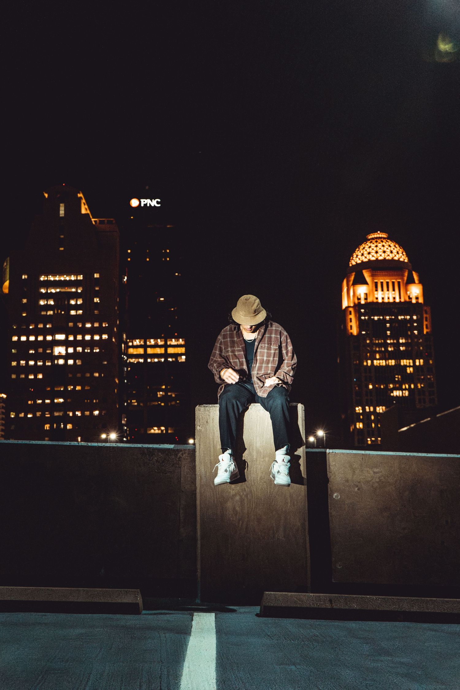 Person in plaid shirt sitting on ledge against illuminated city skyline at night.