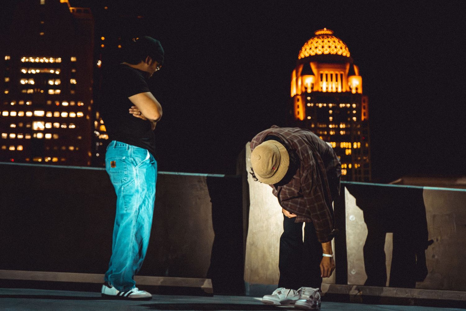 Dramatic nighttime scene with urban buildings and figure silhouetted against glowing cityscape.
