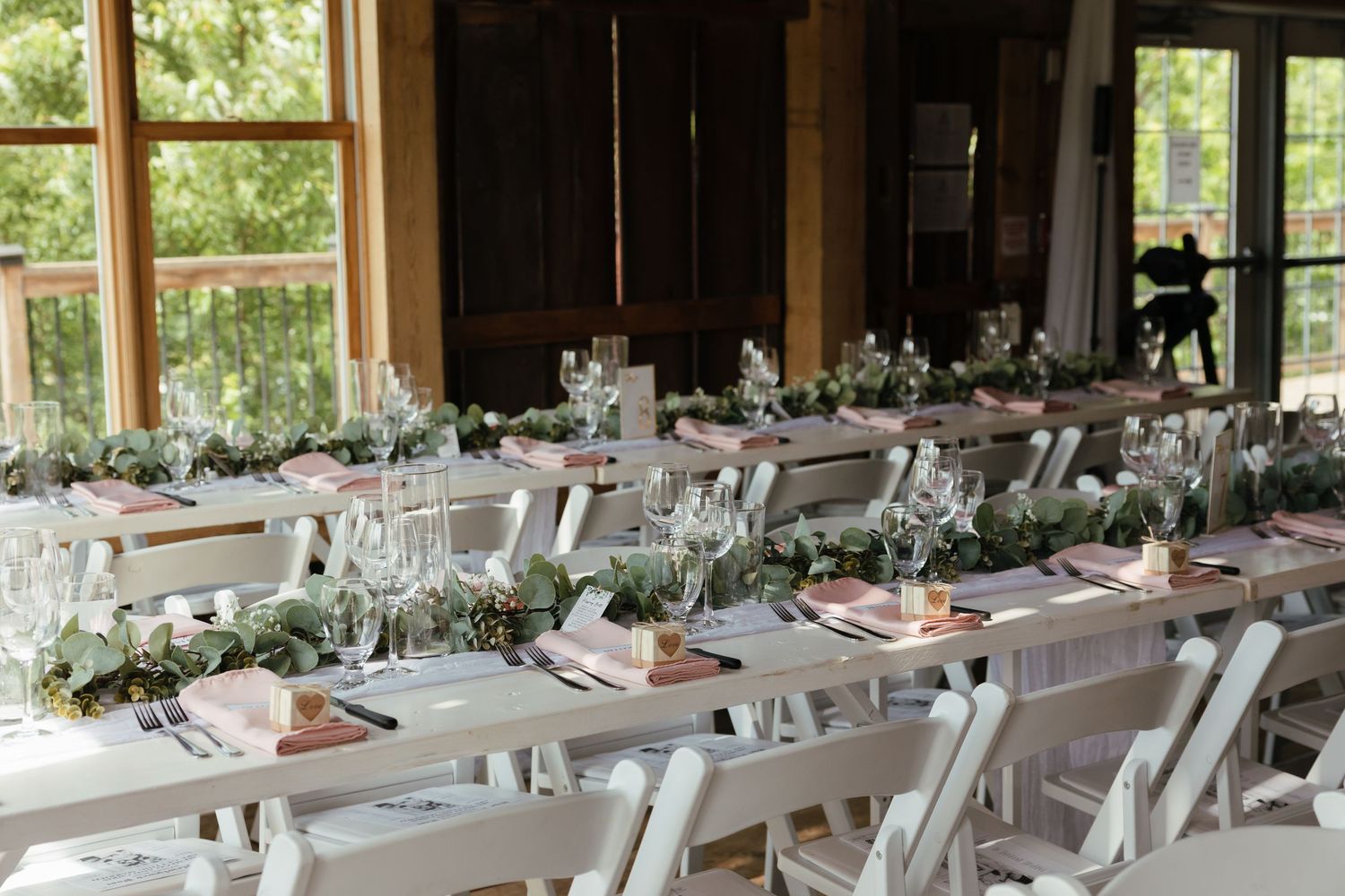 Long banquet tables set for an elegant wedding reception with white linens and greenery centerpieces in rustic venue.