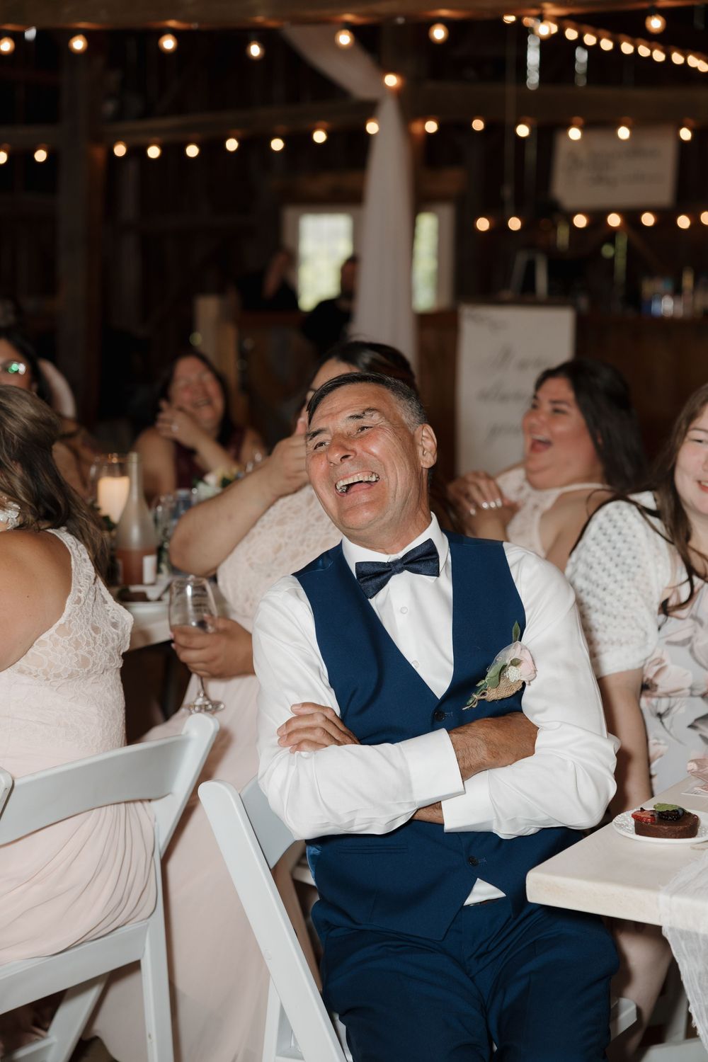 Wedding guests laugh and enjoy themselves at a reception under warm string lights in a romantic venue setting.