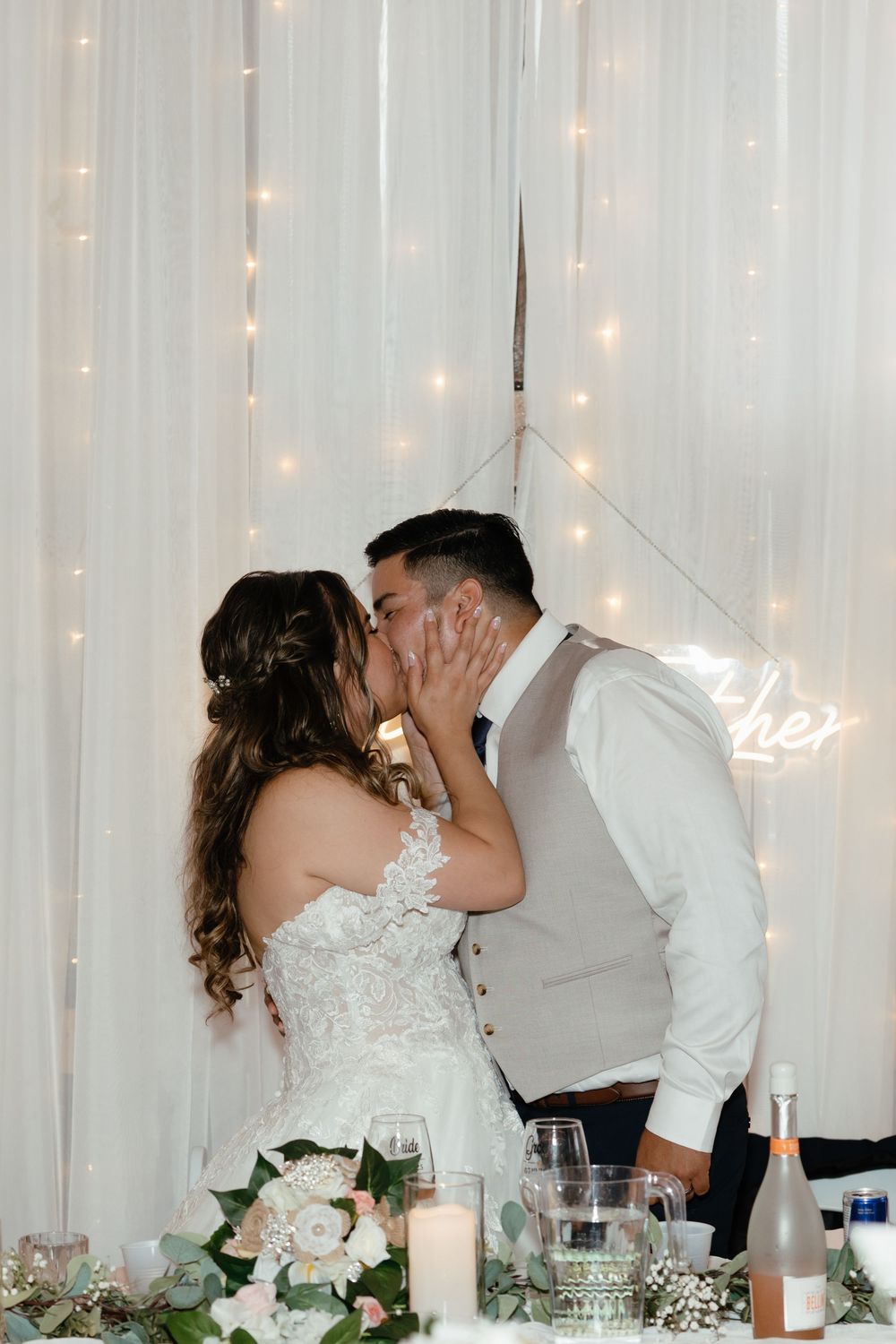 Romantic couple sharing an intimate kiss at their wedding reception with twinkling lights and white decor in background.