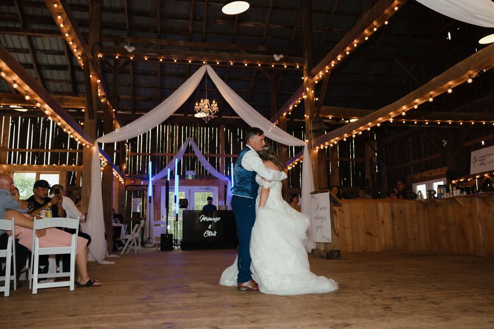 Wedding party dances and celebrates inside a rustic barn venue decorated with string lights and white fabric draping.