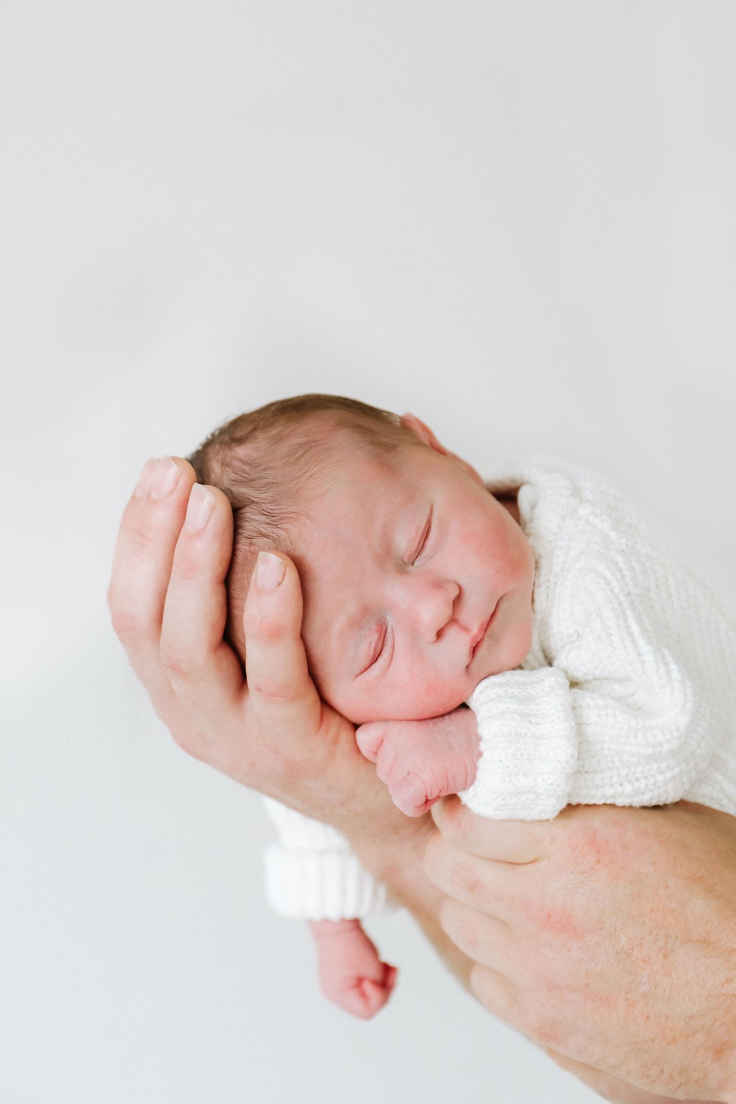 A sleeping newborn baby wearing a white knit sweater rests peacefully in an adult's hands during newborn shoot in Nottingham